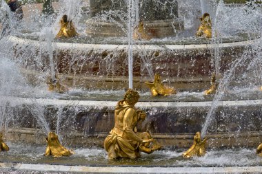 Multi-tiered fountain with golden statues of human figure & frogs spouting water. White water obscures details, bright daylight at Versailles Gardens, Paris.