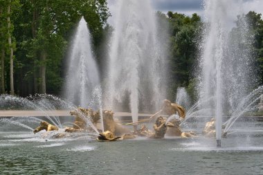 Çeşmedeki altın heykeller ve uzun su püskürtüleri. Ağaçlar arka planda bulutlu bir gökyüzünün altında Versailles Gardens, Paris 'te.