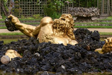 Bir adamın altın heykeli kısmen siyah kayalara, suya, Versailles Gardens, Paris 'teki manikürlü bir bahçenin önüne batar..