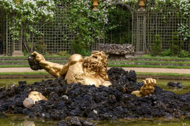 Paris, Versailles Gardens 'da çardağı ve çiçekleri olan bir bahçede, sudaki siyah kayalardan çıkan bir adamın altın heykeli..