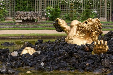 Altın heykel kısmen siyah kaya ve suya batırılmış. Versailles Gardens, Paris 'te arka planda görünen resmi bahçeler.