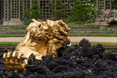 Bahçe ortamında siyah kayalardan çıkan bir adamın altın heykeli. Arka planda Paris 'teki Versailles Gardens' da bir çardak ve manikürlü yeşillik yer alıyor..