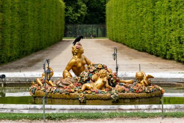 Gilded statue of figures on a floral raft in a pool, with a black bird perched on one figure's head. Hedged path in the background at Versailles Gardens, Paris.