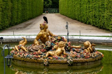 Fountain featuring golden statues of a woman, putti, and flowers with a bird on the woman's head in a garden setting at Versailles Gardens, Paris.