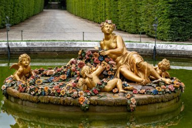 Bir kadının altın heykeli ve çiçeklerle çevrili üç melek Paris 'teki Versailles Gardens' ın arka planında manikürlü bahçe yolu olan bir gölde oturuyor..