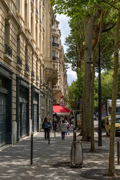 Bej binaların, ağaçların ve park halindeki araçların önünden geçen yayaların olduğu şehir caddesi. Gölgeler kaldırıma düşer.