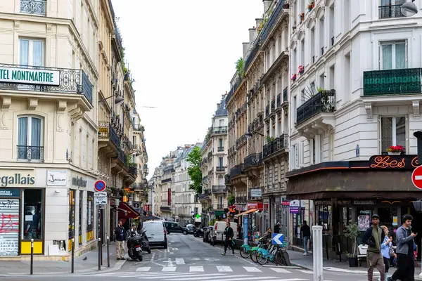 Paris sokak manzarası bulutlu bir günde binaları, insanları, araçları ve dükkanları gösteriyor.