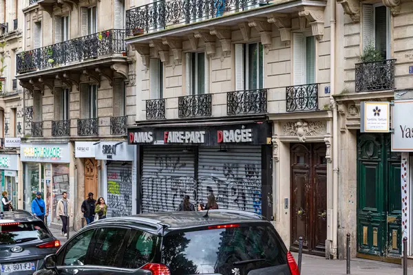 Kaldırımlarda mağazaların, apartmanların, arabaların ve yayaların olduğu bir Paris sokak sahnesi..