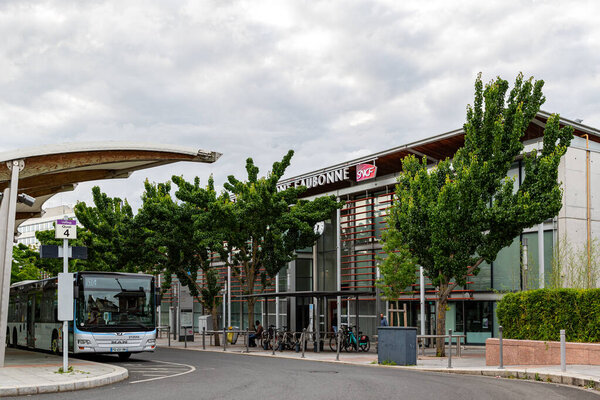 A bus at a station with a train station in the background. Trees and cloudy sky contribute to the outdoor scene.