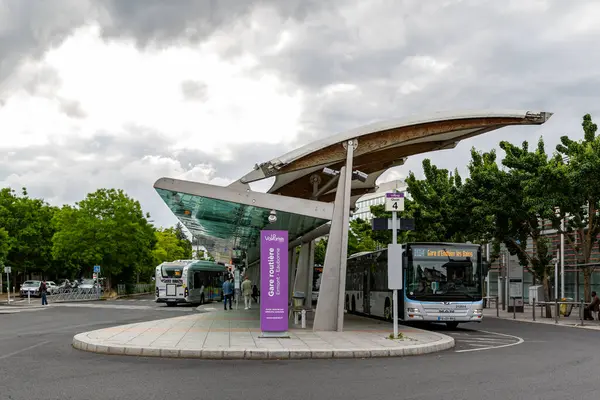 Modern bir barınağı, otobüsleri ve bulutlu bir gökyüzünün altında yeşilliği olan otobüs durağı. Yayaların ve araçların bulunduğu sokak sahnesi.
