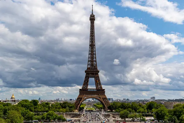 Eyfel Kulesi, şehir manzarası ve yeşil ağaçlarla çerçevelenmiş parçalı bulutlu gökyüzüne karşı dimdik ayakta duruyor..