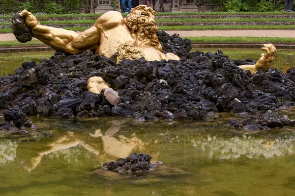 Göldeki siyah kayalardan yükselen altın heykel suya yansıyor. Versailles Gardens, Paris 'te arka planda park ayarları görünür.