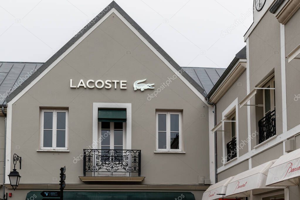 Exterior view of a gray building featuring a Lacoste store sign and windows. The composition is symmetrical at La Vallee Village, France.