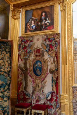 Interior shot of artwork: A painting above a tapestry. Ornate gold details on columns & frames. Two small stools sit in front at Versailles Palace, Paris.