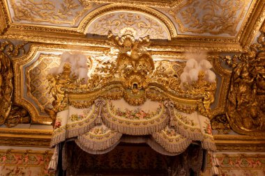Ornate golden canopy bed with floral fabric under a gilded ceiling with detailed carvings and an eagle sculpture. Rich, lavish interior at Versailles Palace, Paris.