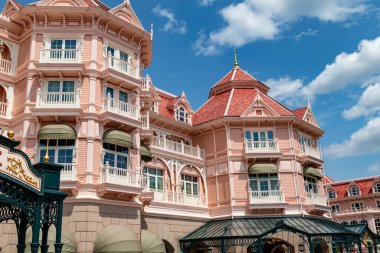 Beyaz çerçeveli, kırmızı çatılı, balkonlu ve yeşil tenteli çok katlı pembe bir binanın dış görüntüsü Disneyland Paris 'te beyaz bulutlu mavi bir gökyüzüne karşı kurulmuş..