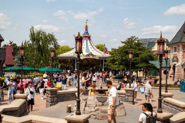 Disneyland Paris 'te bulutlu bir gökyüzü altında kalabalık bir açık hava sahnesi, yürüyen insanlar ve fantezi mimarisi..