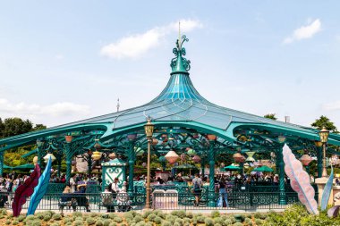 Mavi gökyüzünün altında, Disneyland Paris 'te dağınık bulutlarla dolu, mavi renkli, süslü bir atlıkarınca barınağı insanlarla ve dönen çay fincanlarıyla dolu..