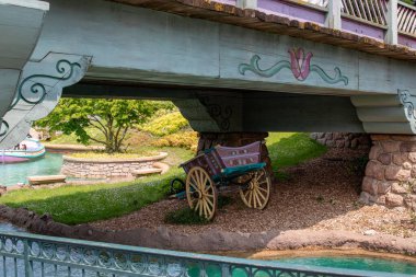 Pastel bir köprünün altında, yemyeşil bir su yolu ve Disneyland Paris 'in arka planında görünür bir tekne bulunan dekoratif bir araba duruyor..