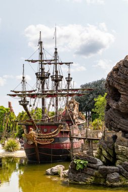 Büyük, dekoratif bir korsan gemisi, detaylı kaya oluşumları ve Paris Disneyland 'de yemyeşil yeşilliklerle dolu yeşil bir gölete kenetlendi..