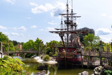 The image shows a docked, red and black pirate ship, surrounded by lush greenery, water, and wooden bridges under a cloudy sky.