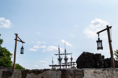 Resim, taş bir duvarın üzerinde bir korsan gemisi silueti gösteriyor. Tahta direkler üzerinde yanan fenerlerle çevrili, Disneyland Paris 'te bulutlu mavi bir gökyüzüne karşı kurulmuş..
