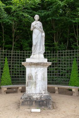 Bahçede sergilenen bir heykel heykeli. Banklar kaideyi kuşatın. Yeşil ağaçlar ve çitler Paris 'teki Versailles Sarayı' nın arka planını oluşturur..