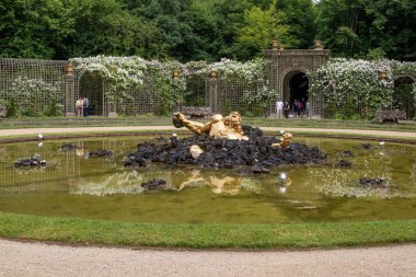 Göldeki koyu kayalardan yükselen bir erkek figürünün süslenmiş altın heykeli. Paris, Versailles Sarayı 'nda arka planda trellises ve yeşillik.
