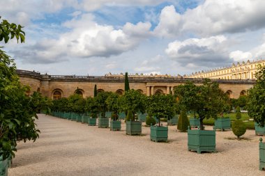 Versailles saray bahçeleri, saksı ağaçları, çakıl yolları ve bulutlu gökyüzünün altında süslü binalar..