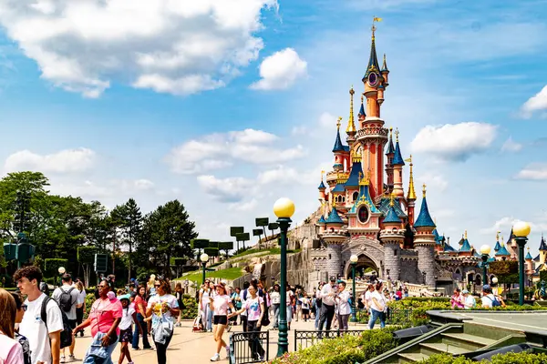 Güneşli bir günde Disneyland Paris 'te, parlak mavi bir gökyüzünün altında tüylü beyaz bulutlarla dolu bir peri masalı kalesinin önündeki kalabalık..