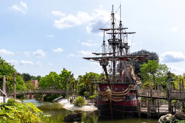 Yeşilliklerle çevrili sudaki korsan gemisi. Arkaplanda ahşap köprü, üzerinde bulutlar olan mavi gökyüzü..