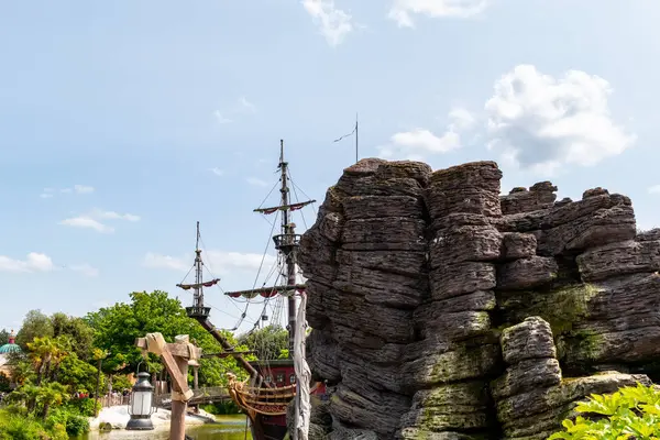 Bir kaya oluşumunun arkasındaki korsan gemisinin manzarası, ağaçlar ve bulutlu mavi gökyüzü Disneyland Paris 'te..