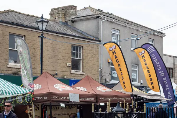 Çadırların altında satıcı tezgahları, renkli reklam bayrakları ve arka planda eski tuğla binalar olan dış pazar sahnesi..