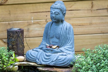 Serin bir Buda heykeli, yeşilliklerle çevrili renkli taşlar taşıyor ve ahşap bir çitin arkasına kuruluyor..