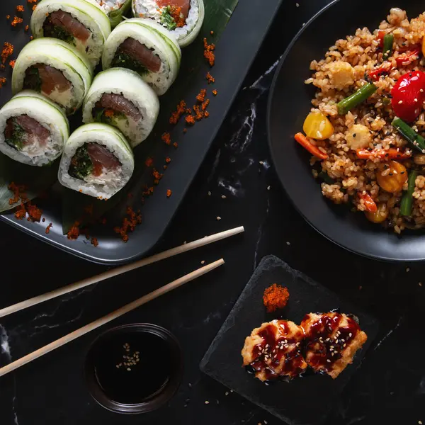 Sebzeli pilav ve siyah tabakta jöleli aperatif ile servis edilen lezzetli salatalıklı suşi ruloları. Koyu bir arka planda modern Asya mutfağı..