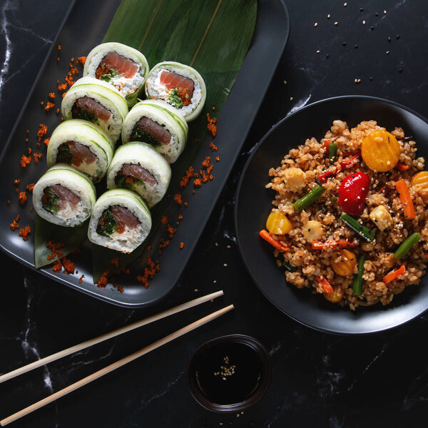 Вкусные суши с огурцом, завернутые в суши, подаются с овощным жареным рисом и глазурованной закуской на черной тарелке. Идеально оформленная современная азиатская кухня на темном фоне.