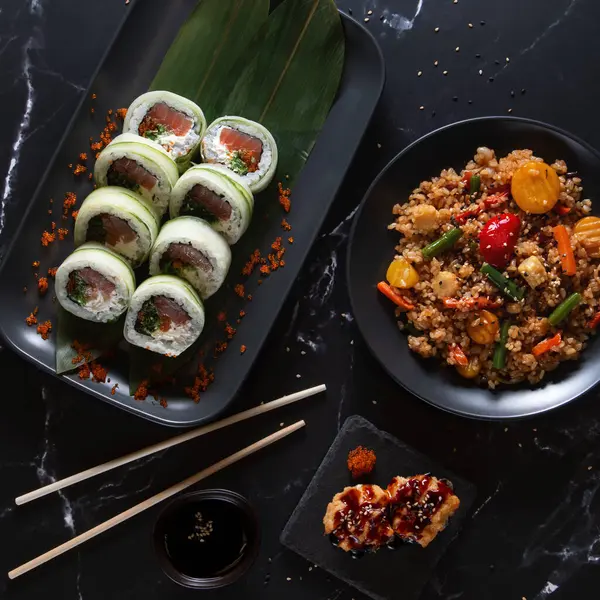 Sebzeli pilav ve siyah tabakta jöleli aperatif ile servis edilen lezzetli salatalıklı suşi ruloları. Koyu bir arka planda modern Asya mutfağı..