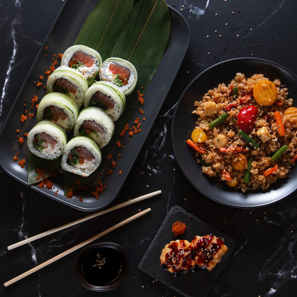 Вкусные суши с огурцом, завернутые в суши, подаются с овощным жареным рисом и глазурованной закуской на черной тарелке. Идеально оформленная современная азиатская кухня на темном фоне.