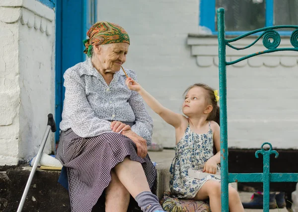 少女と老婦人が老人ホームの階段に腰を下ろしている 女の子はビスケットの藁を食べ 彼女の祖母とそれらを共有します ウクライナの村 祖母と孫娘との家族関係 — ストック写真