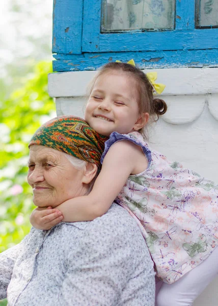 Küçük bir kız yaşlı büyükannesine sıkıca sarılıyor ve gülüyor. Ukrayna köyü. Büyükannemle torunum arasındaki ilişki.