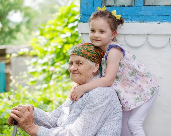 Küçük bir kız yaşlı büyükannesine sıkıca sarılıyor ve gülüyor. Ukrayna köyü. Büyükannemle torunum arasındaki ilişki.