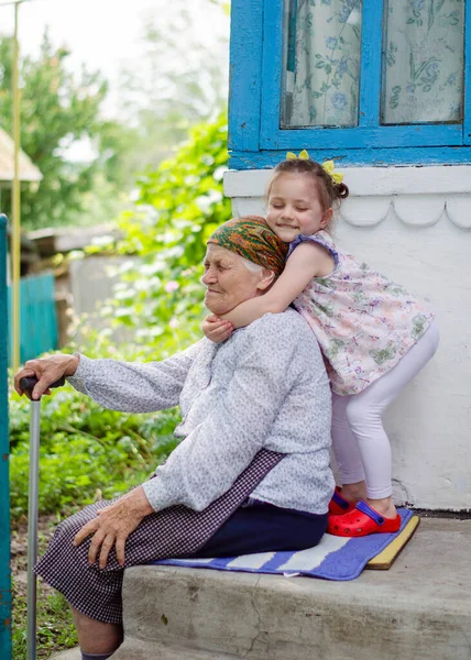 Küçük bir kız yaşlı büyükannesine sıkıca sarılıyor ve gülüyor. Ukrayna köyü. Büyükannemle torunum arasındaki ilişki.