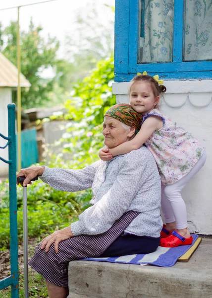 Küçük bir kız yaşlı büyükannesine sıkıca sarılıyor ve gülüyor. Ukrayna köyü. Büyükannemle torunum arasındaki ilişki.