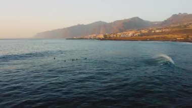 Tenerife adasının batı kıyısındaki turistik Los Gigantes tatil beldesinin üzerinde yükselen dumanlı dağ silsilesiyle. Güçlü rüzgar, koyu mavi okyanusta büyük dalgalara neden olur. Gün batımında kumsalı yıkar.