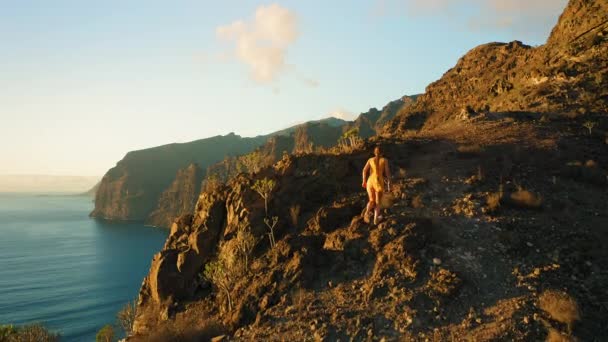 若い女性の崖の上に海を見下ろす上を歩く 少女は山の自然の美しさを観察登山 美しい夕日の空 空中ドローンビュー — ストック動画