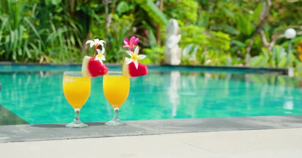 两瓶有花的黄色鸡尾酒果汁紧靠着果肉端站着 游泳池蓝色水底 翠绿茂密的热带雨林棕榈树度假 慢动作视频 — 图库视频影像