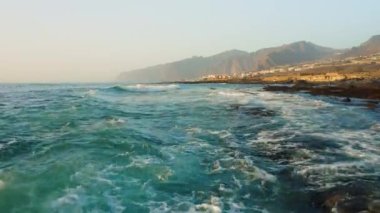 Fırtınalı havada insansız hava aracı okyanus yüzeyinde alçaktan uçuyor. Deniz dalgaları köpürüyor. Kaya kıyısı. Arka planda Los Gigantes uçurumları var. Tenerife. Turkuaz Atlantik sularının güzel manzarası. Kanarya