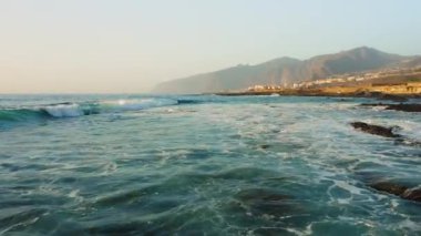 Fırtınalı havada insansız hava aracı okyanus yüzeyinde alçaktan uçuyor. Deniz dalgaları köpürüyor. Kaya kıyısı. Arka planda Los Gigantes uçurumları var. Tenerife. Turkuaz Atlantik sularının güzel manzarası. Kanarya