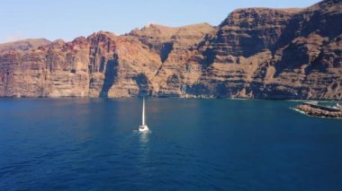 Büyük yüksek dağlar ve derin mavi gök mavisi okyanus sularında sürüklenen yalnız beyaz bir yat. Parlak bir öğleden sonra küçük bir teknenin havadan görüntüsü. Tenerife Kanarya Adası İspanya. Doğa manzarası.