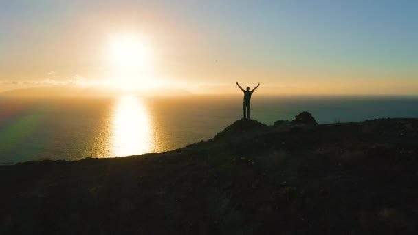 カラフルな夕日の上に立って手を挙げる旅行男がいる 海の背景に大きな座って太陽とオレンジの空を見て男のシルエット 山の風景 冒険旅行 — ストック動画